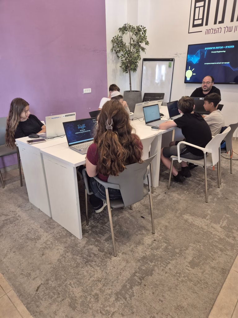 קבוצת ילדים יושבת סביב שולחן לבן, כל אחד עובד על מחשב נייד, ומדריך מבוגר עומד לידם. ברקע מוצגת מצגת עם טקסט בעברית. חדר החוגים בפרדס חנה צבוע בסגול ולבן.
