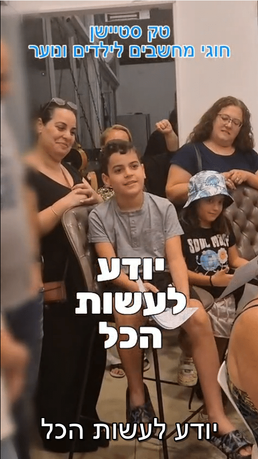 ילד יושב ומחייך בכיתה במהלך חוג מחשבים, מוקף במבוגרים וילדים. חלק מהאנשים מוחאים כפיים ומחייכים. על התמונה מוטבע טקסט בעברית.