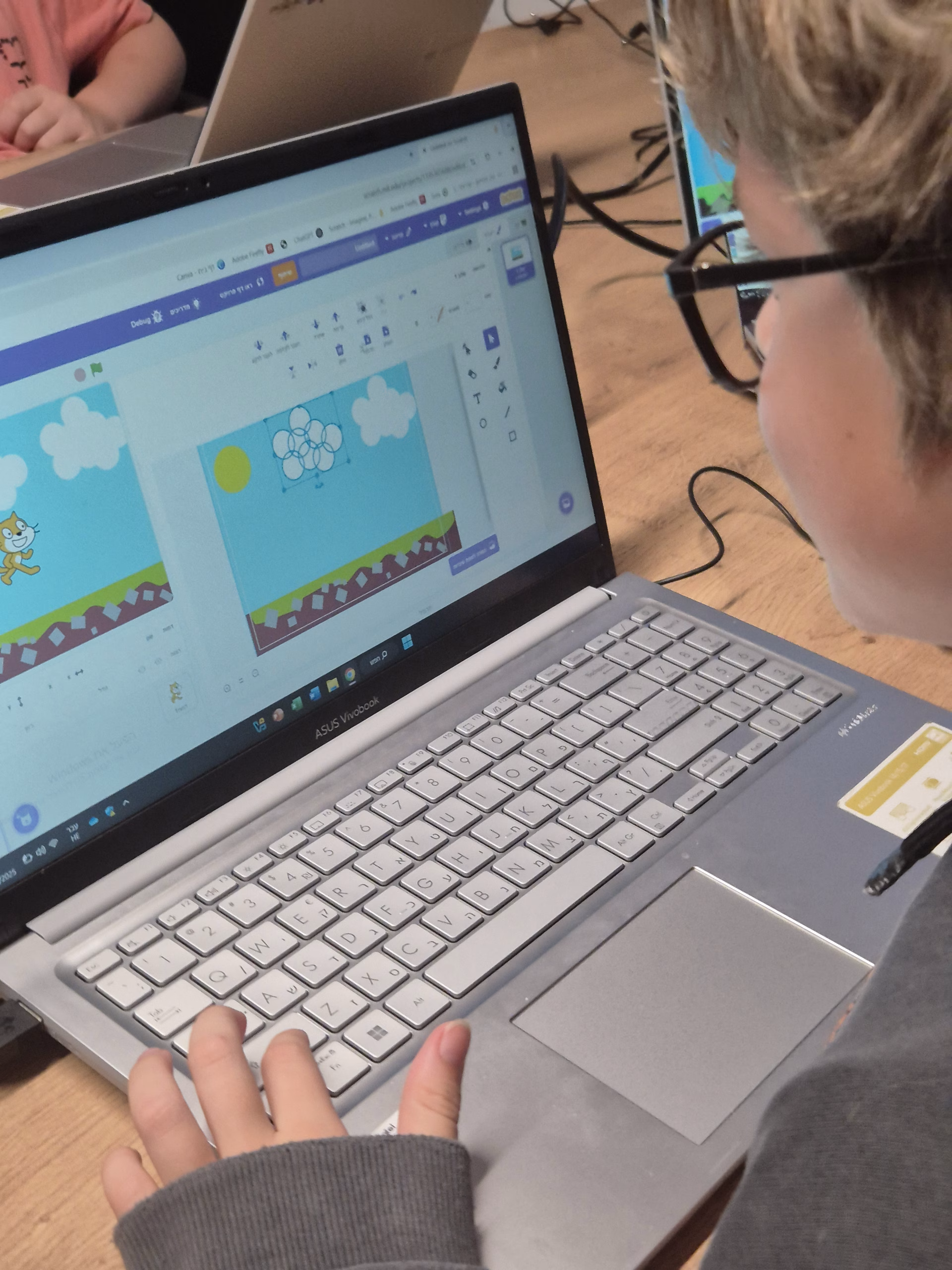 ילד המרכיב משקפיים משתמש במחשב נייד כדי ליצור אנימציה באמצעות Scratch בחוג מחשבים, המציגה חתול ועננים על רקע מצויר. ברקע נראים מחשבים ניידים נוספים ואנשים.