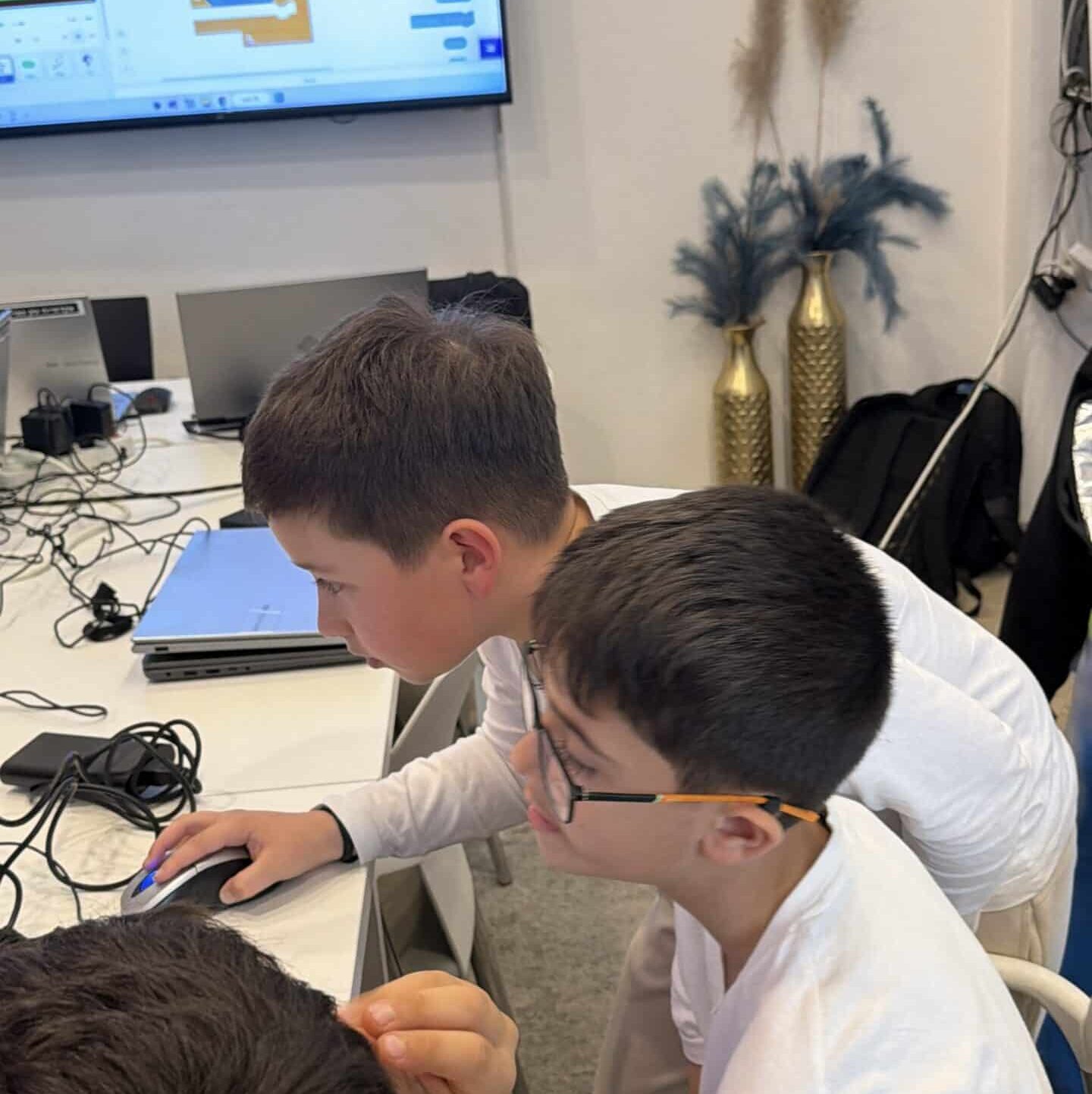 שלושה נערים בחולצות לבנות יושבים יחד ליד שולחן מחשב במהלך חוג, ומביטים במסך המציג תוכנת תכנות. על השולחן מונחים כבלים ומחשב נייד, וברקע נראית מסך עם תרשימים וקוד.