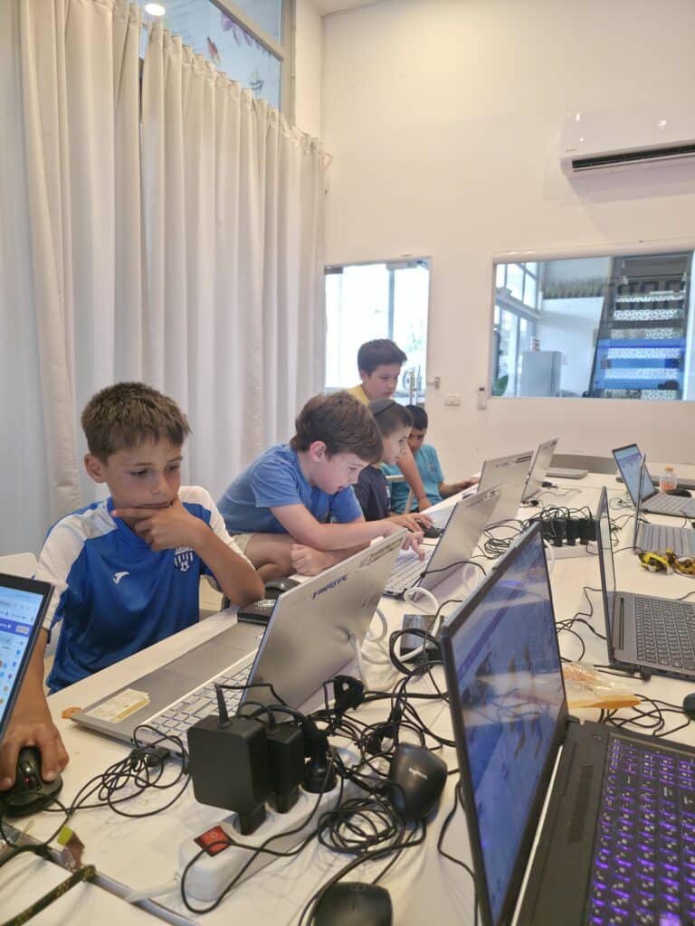 ארבעה נערים יושבים ליד שולחן עם מחשבים ניידים, מרוכזים במסכים שלהם. החדר מואר, עם קירות ווילונות לבנים, וכמה כבלים ומכשירים פזורים על השולחן.