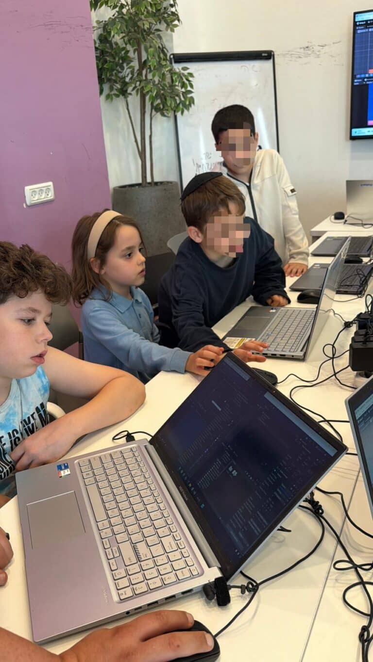 ארבעה ילדים יושבים סביב שולחן ומשתמשים במחשבים ניידים, כשהם מרוכזים במסכים שלהם. על המסכים נראות תוכנות לתכנות או לקוד. ברקע נראים עציץ ולוח מחיק. חלק מהפנים מטושטשות מטעמי פרטיות.
