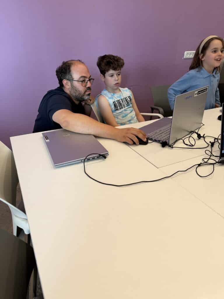 גבר יושב ליד שני ילדים ליד שולחן לבן, ועוזר לאחד מהם עם מחשב נייד. נראה שהשלושה מרוכזים, והקיר ברקע צבוע בסגול.