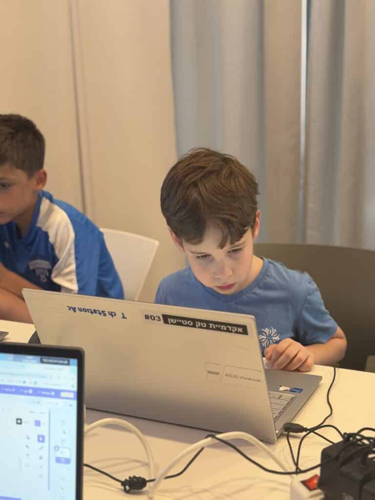 ילד לבוש בחולצה כחולה יושב ליד שולחן ומביט במסך המחשב הנייד, בעוד ילד אחר יושב בסמוך אליו ומשתמש גם הוא במחשב. התמונה נראית כמו כיתה או סדנה.