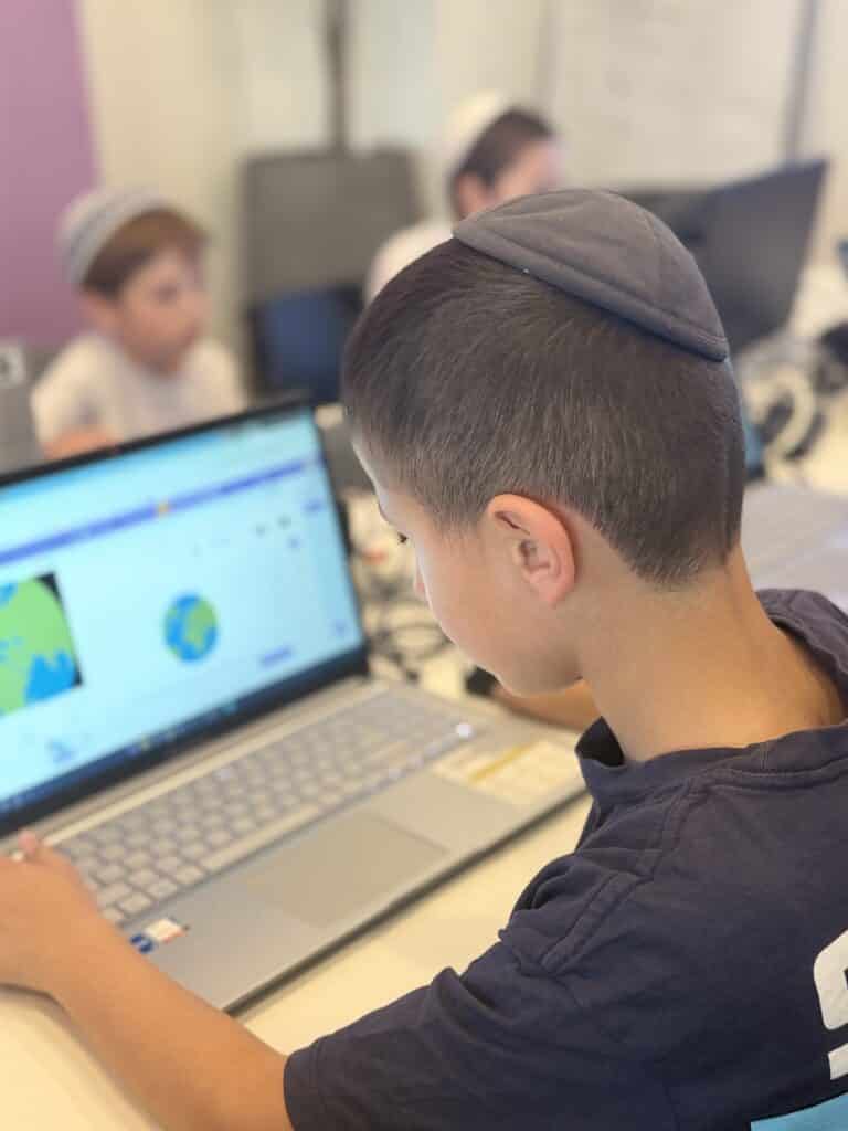 ילד חובש כיפה יושב מול מחשב נייד, מתבונן במסך שעליו מופיעה איור דיגיטלי של גלובוס. ברקע נראים ילדים נוספים היושבים מול מחשבים בכיתה.