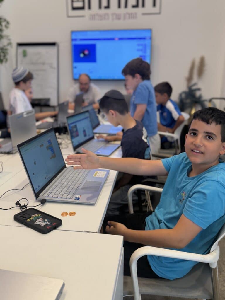 ילד מחייך יושב ליד שולחן עם מחשב נייד, ומצביע על המסך. סביבו, ילדים אחרים עובדים גם הם על מחשבים ניידים בכיתה, כשברקע נראים מורה ומסך גדול.