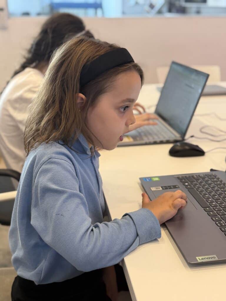 ילדה צעירה לבושה בחולצה כחולה וברצועת ראש שחורה יושבת ליד שולחן ומרוכזת בעבודה על מחשב נייד, כשברקע אדם נוסף עובד על מחשב נייד.