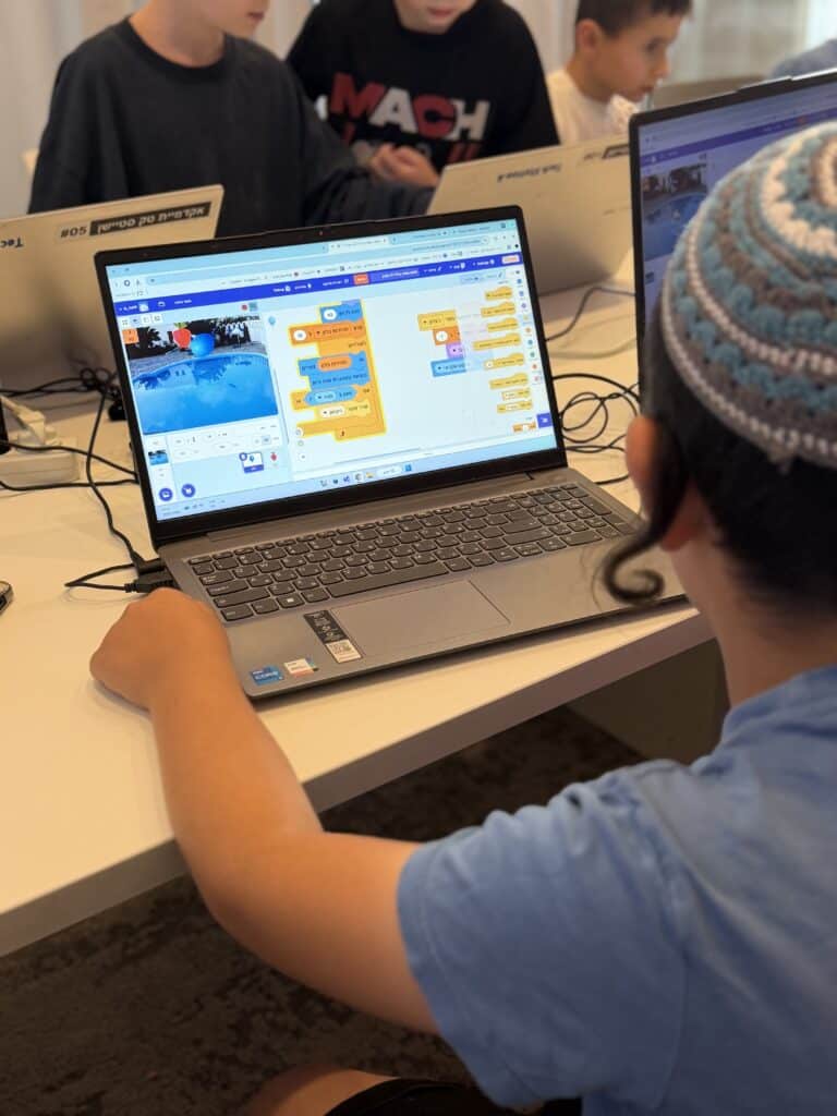 ילד לבוש בחולצה כחולה וקיפּה מעוטרת עובד על תכנות פרויקט ב-Scratch במחשב נייד. ברקע, סביב השולחן, נראים גם ילדים אחרים עם מחשבים ניידים.