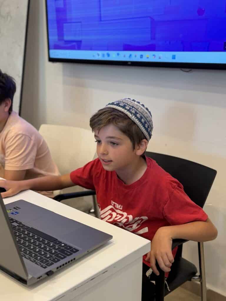 נער לבוש בחולצה אדומה וכיפה סרוגה בכחול-לבן יושב ליד שולחן ומשתמש במחשב נייד. ברקע נראית מסך גדול עם תצוגה כחולה. אדם נוסף נראה חלקית לצדו.