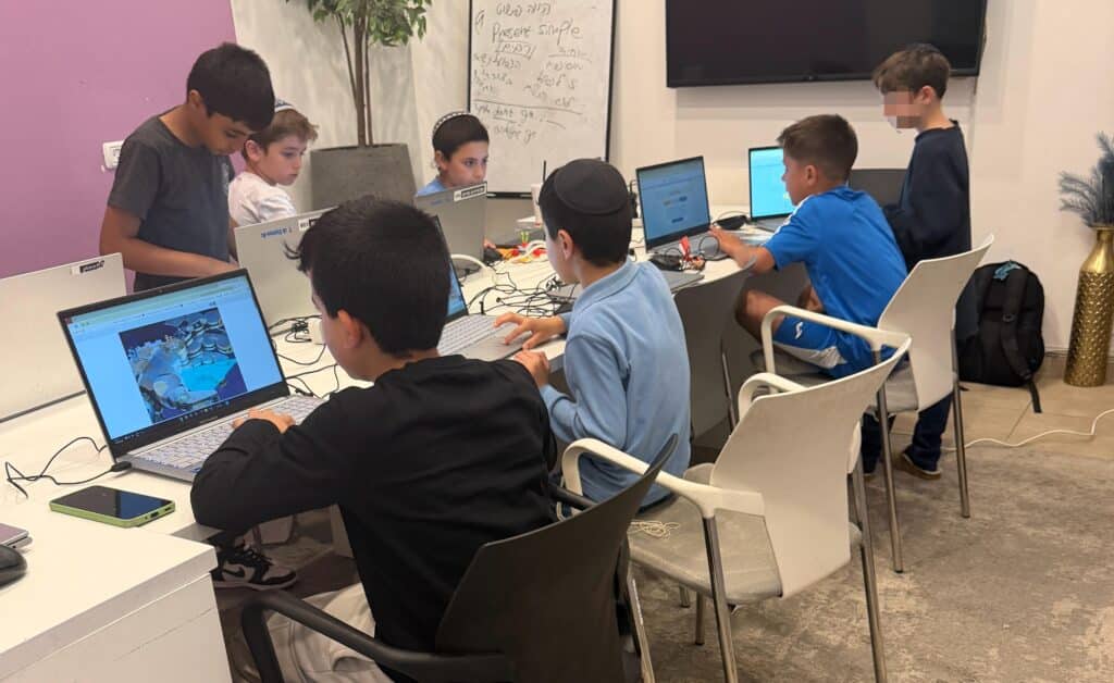 קבוצת ילדים יושבת סביב שולחן עם מחשבים ניידים, ונראית מרוכזת במסכים שלה. חלקם עוסקים בתכנות או בפיתוח משחקים לילדים, בעוד שאחרים משוחחים ביניהם. המקום נראה כמו כיתה או סדנת טכנולוגיה.