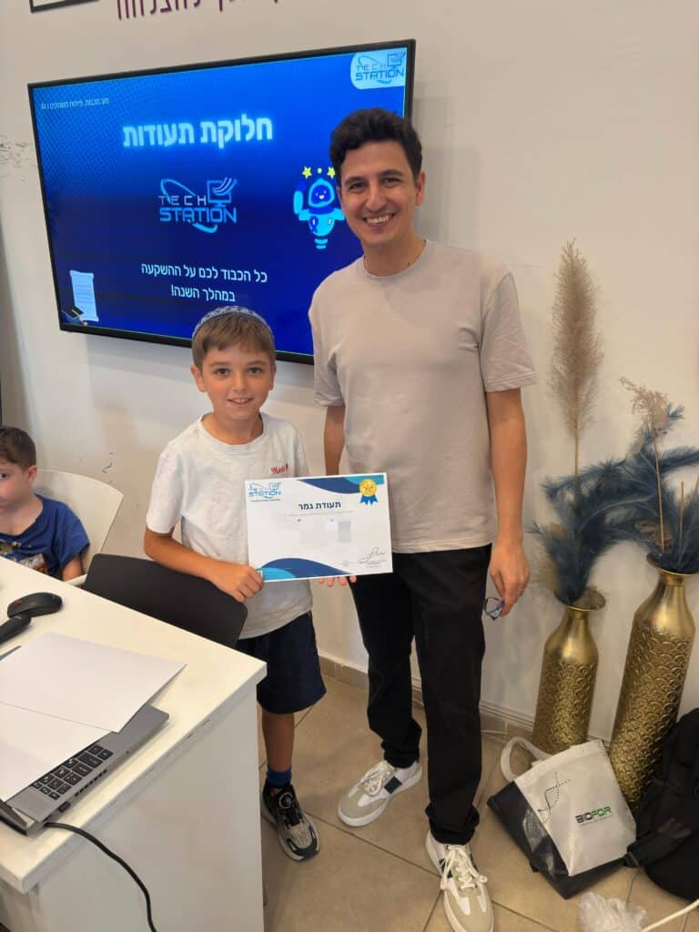 ילד מחייך עומד לצד מבוגר, אוחז בתעודה מהחוג שלו. הם נמצאים בתוך חדר, ליד שולחן שעליו מחשב נייד. ברקע, על המסך, מופיעים טקסט בעברית ואיור של רקטה. ילד נוסף יושב בסמוך.