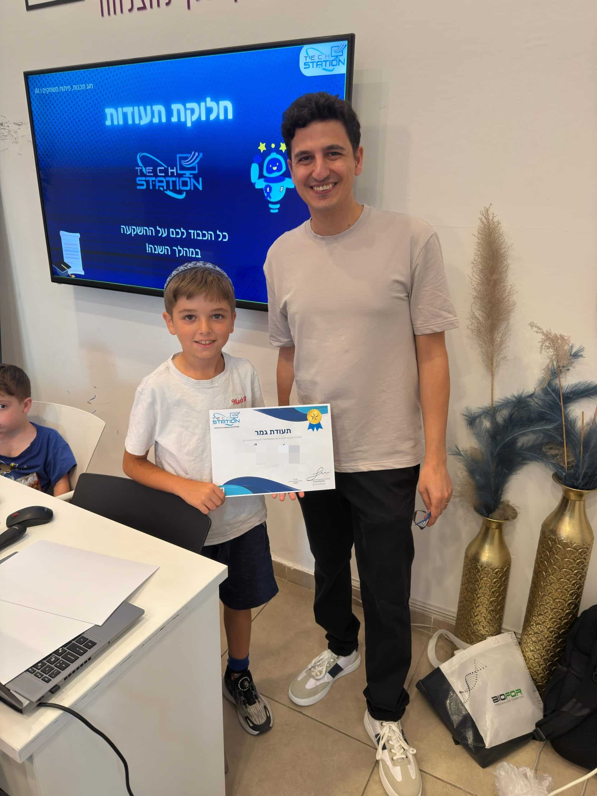 ילד מחייך עומד לצד מבוגר, אוחז בתעודה מהחוג שלו. הם נמצאים בתוך חדר, ליד שולחן שעליו מחשב נייד. ברקע, על המסך, מופיעים טקסט בעברית ואיור של רקטה. ילד נוסף יושב בסמוך.