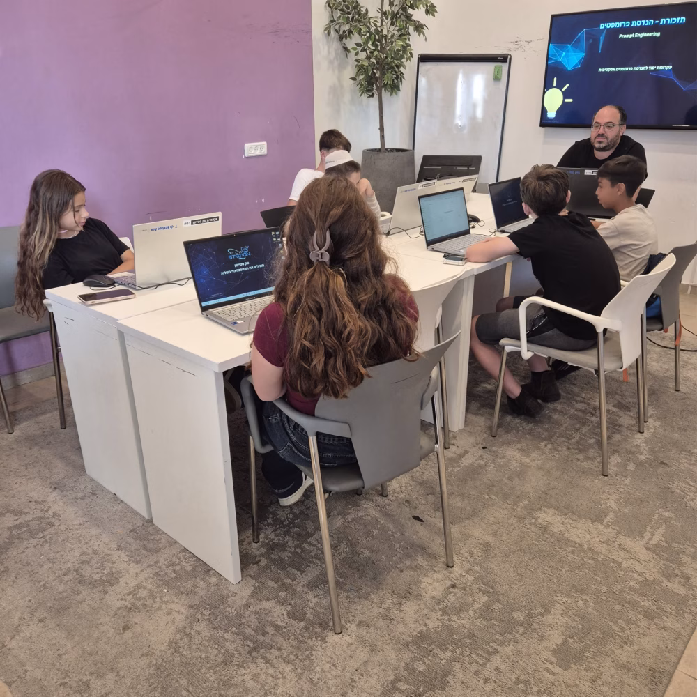 קבוצת ילדים יושבת סביב שולחן לבן, כל אחד עובד על מחשב נייד, ומדריך מבוגר עומד לידם. ברקע מוצגת מצגת עם טקסט בעברית. חדר החוגים בפרדס חנה צבוע בסגול ולבן.